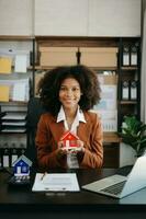 Young real estate agent worker working with laptop and tablet at table in office and small house beside it. photo