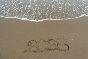 New Year 2026 written in the sand on a beach with sea wave background. photo