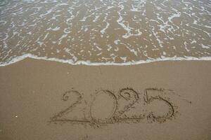 New Year 2025 written in the sand on a beach with sea wave background. photo