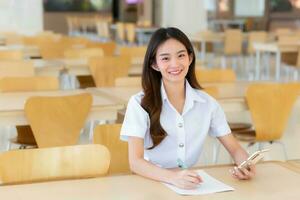 Young Asian woman student in uniform using smartphone and writing something about work. There are documents on table her face with smiling in working to search information for study at university. photo