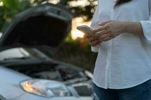 breakdown, broken car, repair. Woman uses mobile phone check insurance premiums through application due to car accident . Find garage to get car fixed during country tour, waiting for help, emergency. photo