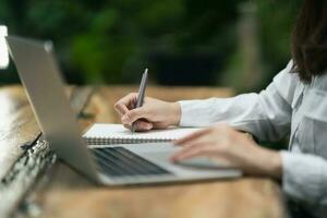 woman in smart casual wear writing on notebook and working on laptop while sitting in creative office or cafe. young girl working with laptop on the wood table in the cafe photo