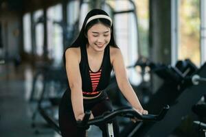 Asian sportswoman exercising on a bicycle in the gym, determination to cardio lose weight, makes her healthy. exercise bike woman fitness sport concept. photo