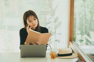 Asian freelance woman smiling using pen making notices her notebook and working on laptop on table at home. Entrepreneur woman working for her business at cafe. Business work at coffee shop concept photo