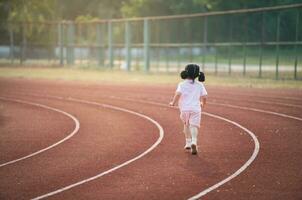 espalda lado. bebé asiático niña correr trotar a corriendo pista, campo a estadio. pequeño niña corriendo puesta de sol contento bebé niña sonriente. pequeño niña corriendo a puesta de sol. linda bebé niña corriendo a corriendo pista. foto