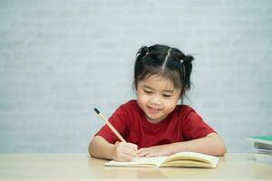 Asian baby girl wearing a red shirt write notes in notebook and reading book to study online on wood table desk in living room at home. Education learning online from home concept. photo