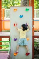 linda asiático niña sonrisa alpinismo escalera y jugar en colegio o jardín de infancia yarda o patio de juegos. sano verano actividad para niños. pequeño asiático niña gracioso contento. niño jugando en al aire libre patio de juegos. foto