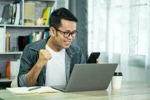 freelance man in eyewear working with laptop and mobile successful cheerful, sitting at wooden table. Pleasant happy man communicating in social network, conferrence vdo call. photo