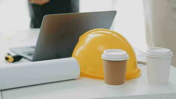 Team of construction workers wearing protective helmets and vests discussing project details with executive supervisor standing at table with blueprints, tools and laptop on it video