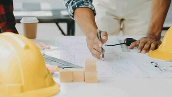 Team of construction workers wearing protective helmets and vests discussing project details with executive supervisor standing at table with blueprints, tools and laptop on it video