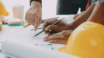 Team of construction workers wearing protective helmets and vests discussing project details with executive supervisor standing at table with blueprints, tools and laptop on it video