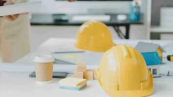 Team of construction workers wearing protective helmets and vests discussing project details with executive supervisor standing at table with blueprints, tools and laptop on it video