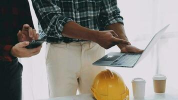 Team of construction workers wearing protective helmets and vests discussing project details with executive supervisor standing at table with blueprints, tools and laptop on it video