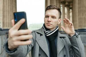 Handsome young man in coat walking on street holds mobile phone and shoots photo