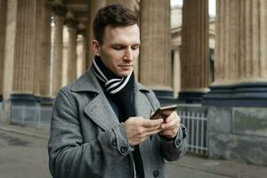 Smiling young man in gray coat is holding scroll text messages in his mobile photo