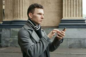 Portrait caucasian young man using his mobile phone in gray coat stands photo