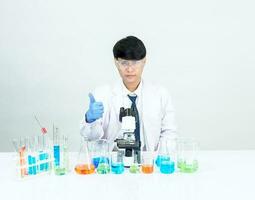 Asian male student scientist Wearing a doctor's gown in the laboratory, looking at the results through a microscope. on a table in a scientific research laboratory with test tubes on a white table photo