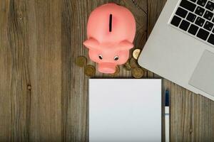 Blank sheet of paper and pink piggy bank close to the laptop. photo