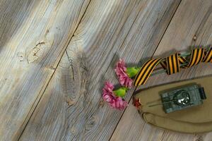 Two pink carnations, Saint George ribbon, tank, and military cap on a wooden surface. photo