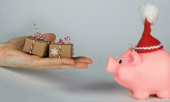 Pink piglet in a Santa Claus hat and human hand with Christmas presents in front of it. photo