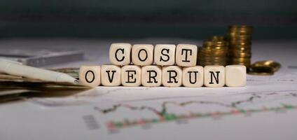 Words COST OVERRUN  composed of wooden letter. Stacks of coins in the background. photo
