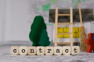 Word  CONTRACT composed of wooden letter. Small paper house, wooden trees in the background. photo