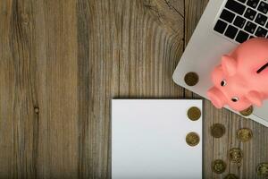 Blank sheet of paper and pink piggy bank close to the laptop. photo