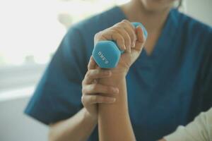 Old woman training with physiotherapist using dumbbells at home. Therapist assisting senior woman with exercises in nursing home. Elderly patient using dumbbells with outstretched arms. photo