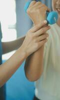 Old woman training with physiotherapist using dumbbells at home. Therapist assisting senior woman with exercises in nursing home. Elderly patient using dumbbells with outstretched arms. photo