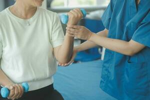 Old woman training with physiotherapist using dumbbells at home. Therapist assisting senior woman with exercises in nursing home. Elderly patient using dumbbells with outstretched arms. photo