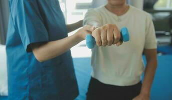 Old woman training with physiotherapist using dumbbells at home. Therapist assisting senior woman with exercises in nursing home. Elderly patient using dumbbells with outstretched arms. photo