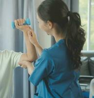 Old woman training with physiotherapist using dumbbells at home. Therapist assisting senior woman with exercises in nursing home. Elderly patient using dumbbells with outstretched arms. photo