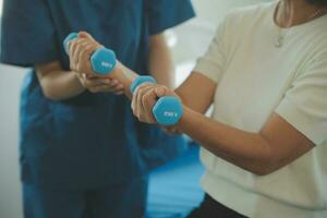 Old woman training with physiotherapist using dumbbells at home. Therapist assisting senior woman with exercises in nursing home. Elderly patient using dumbbells with outstretched arms. photo