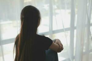 pensativo niña sentado en umbral abrazando rodillas mirando a ventana, triste Deprimido adolescente gasto hora solo a hogar, joven trastornado pensativo mujer sensación solitario o frustrado pensando acerca de problemas foto