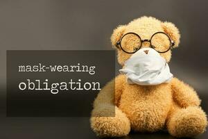 Words MASK-WEARING OBLIGATION. Plush teddy bear in a medical mask in the background photo