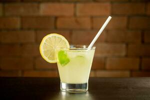 Glass of lime caipirinha drink on top of table in front of a brick wall with text space photo