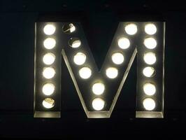 Light bulb glowing letter alphabet character M font. Front view illuminated capital symbol on black background. photo