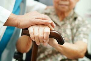 Doctor help Asian elderly disability woman patient holding walking stick in wrinkled hand at hospital. photo