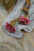 Two pink carnations, Saint George ribbon and military cap on a wooden surface. photo