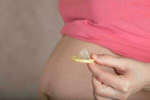 joven embarazada mujer mantiene un condón cerca a su barriga. foto