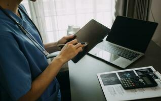 Serious female doctor using laptop and writing notes in medical journal sitting at desk. Young woman professional medic physician wearing white coat and stethoscope working on computer at workplace. photo