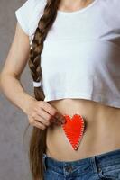 Young lady dressed in jeans shows a heart close to the belly. Closeup photo