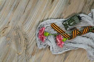 Two pink carnations, Saint George ribbon and military tank on a wooden surface. photo