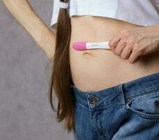 Young lady  dressed in jeans keeps pregnancy test close to the belly. Closeup photo