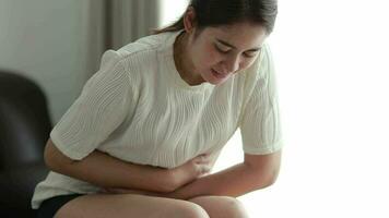 joven asiático mujer estómago dolor sentado en cama a hogar, salud problema inflamación en cuerpo, período ciclo día de mensual, menstruación concepto. video