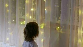 Little boy in christmas pajamas decorating curtain with christmas light beside the christmas tree video