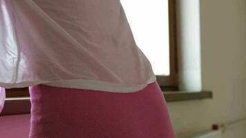 Young woman in a bright pink dress and white cardigan stand beside the window looking outside observing with a smile video