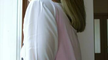 Young woman in a bright pink dress and white cardigan stand beside the window looking outside observing with a smile video