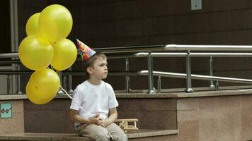 Sad boy of seven years old with birthday balloons playing with wooden airplane, waiting for someone. Boy has a birthday paper hat on his head video