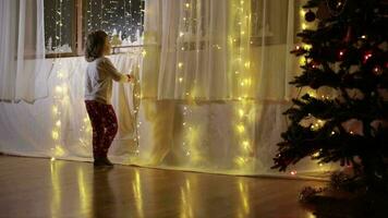 pequeño chico en Navidad pijama decorando cortina con Navidad ligero junto a el Navidad árbol video